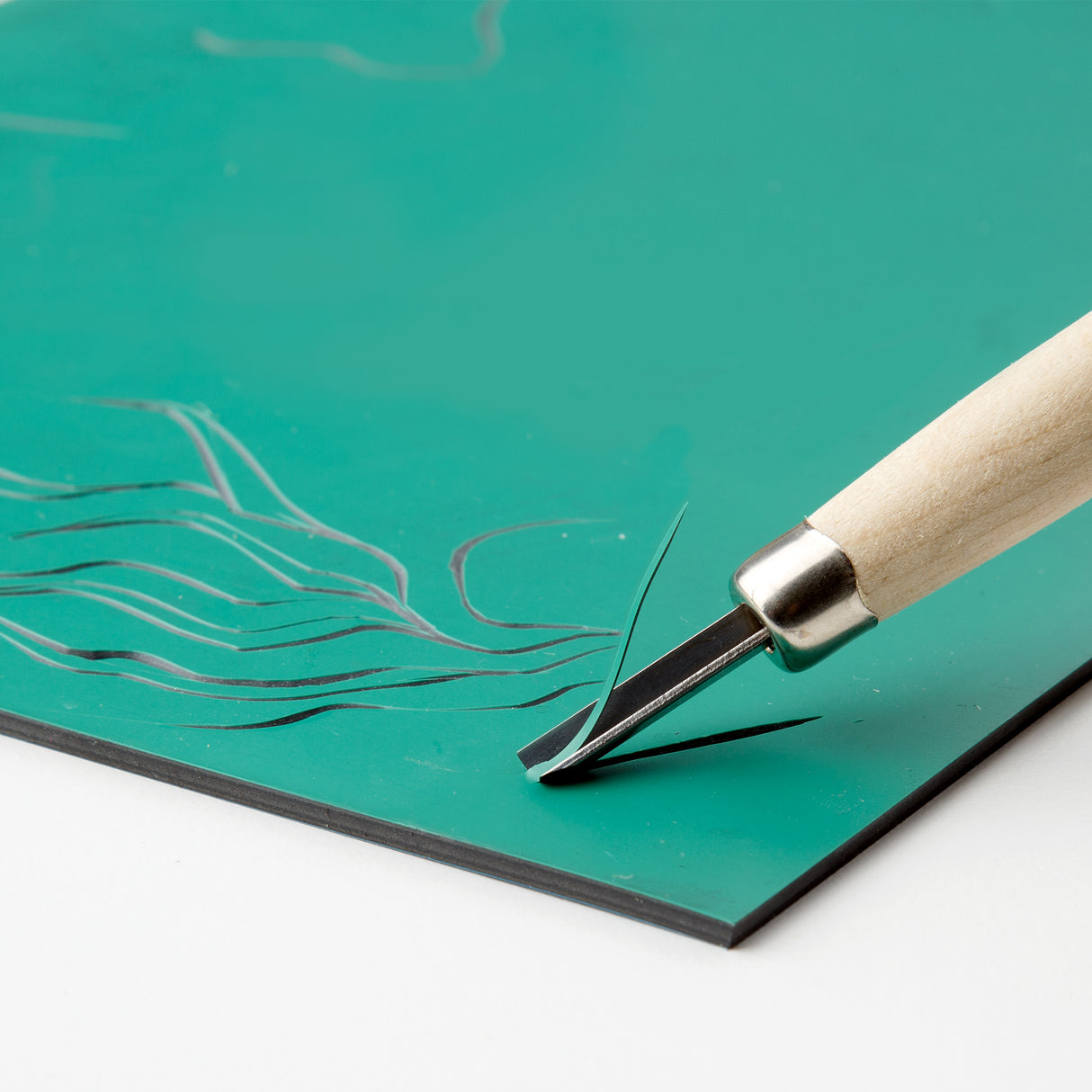 Close-up photograph of a carving tool cutting into a green Lino Tile Vinyl Double Sided sheet. The sharp blade lifts a thin curled shaving as smooth lines are carved into the surface, demonstrating controlled cutting and clean mark making.