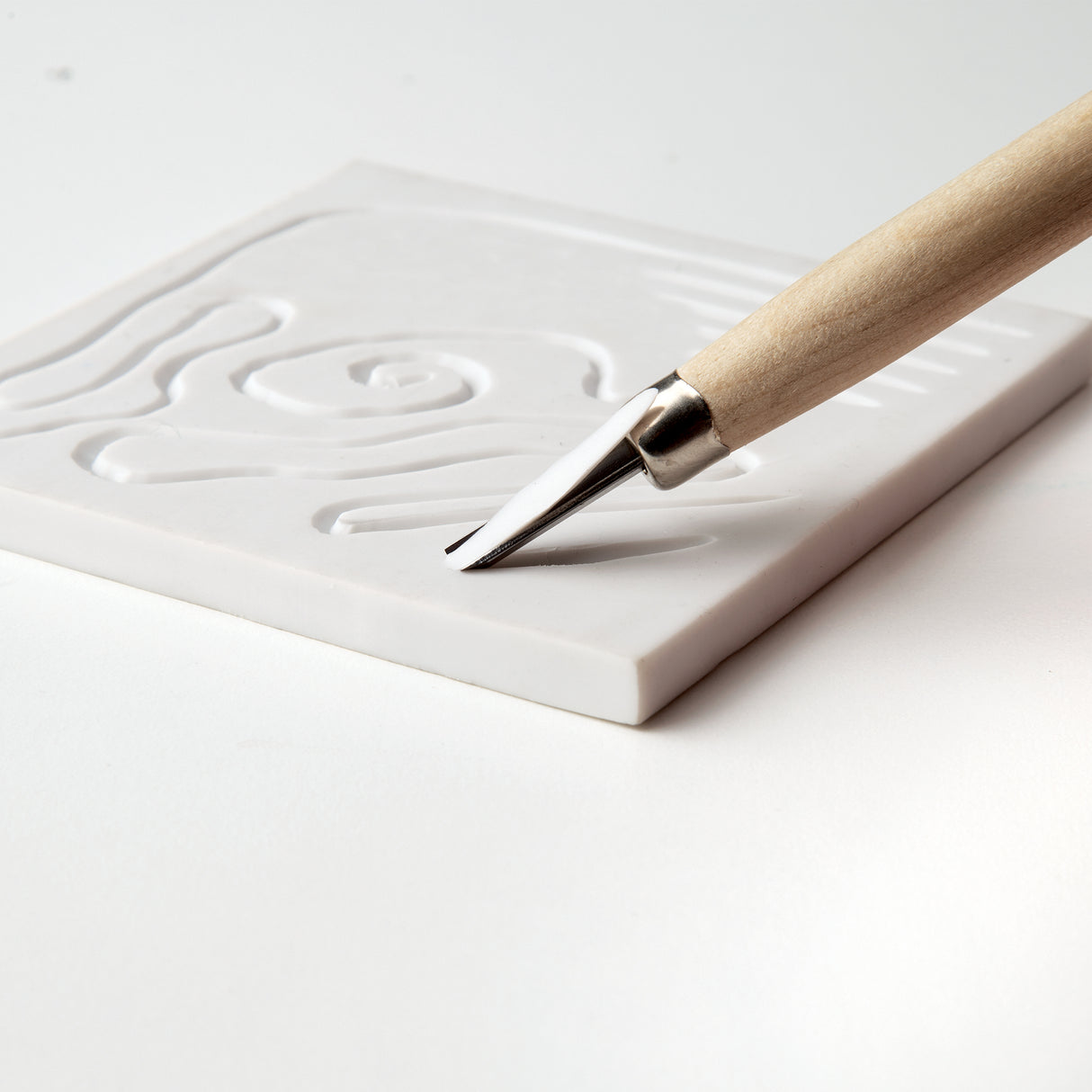 Close-up of a wooden-handled carving tool cutting into a white soft cut block. The metal blade creates smooth recessed lines forming curved shapes. The block has a matte surface and straight, clean edges, resting on a white background.
