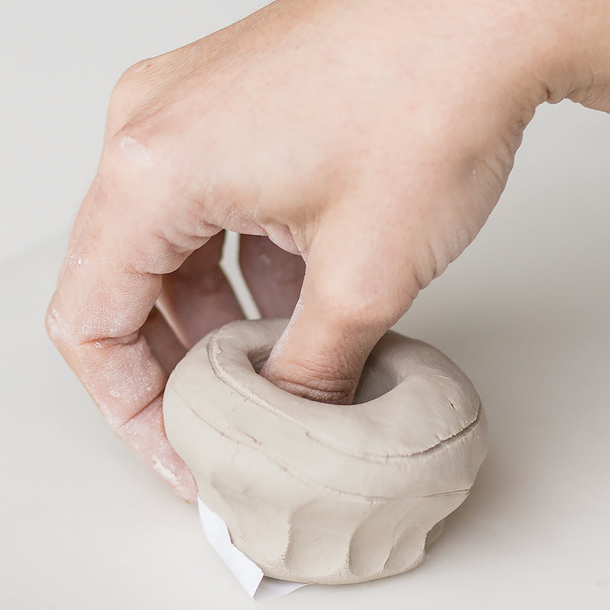 A hand pressing a wooden tool into a cream-coloured clay coil pot with a rounded base and wide opening. The clay vessel sits on white paper, showing the malleable texture of Zart Oz Clay being shaped and smoothed during the handbuilding process.
