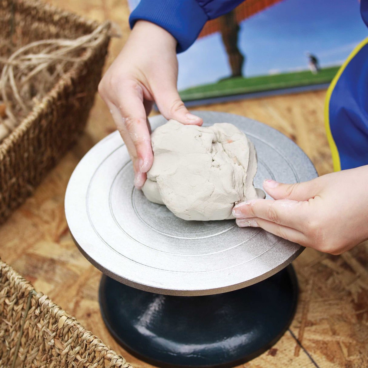 Child's hands working with grey clay on an 18cm Turning Wheel featuring concentric circle guides on a white surface with black base. Woven basket and colourful classroom artwork visible in background, demonstrating hands-on pottery and sculpting techniques in an educational setting.