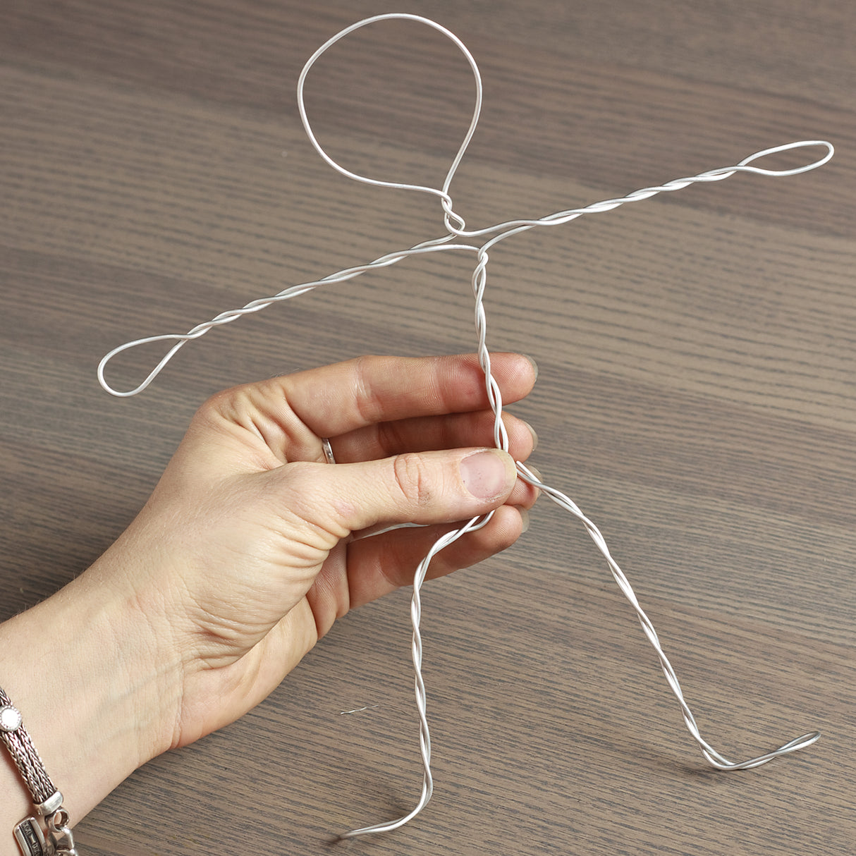 Hand holding pliable Armature Wire twisted into a simple stick-figure shape, demonstrating flexibility and ease of bending. The silver wire is partially twisted to form arms, legs and a looped head, shown against a wooden tabletop background.