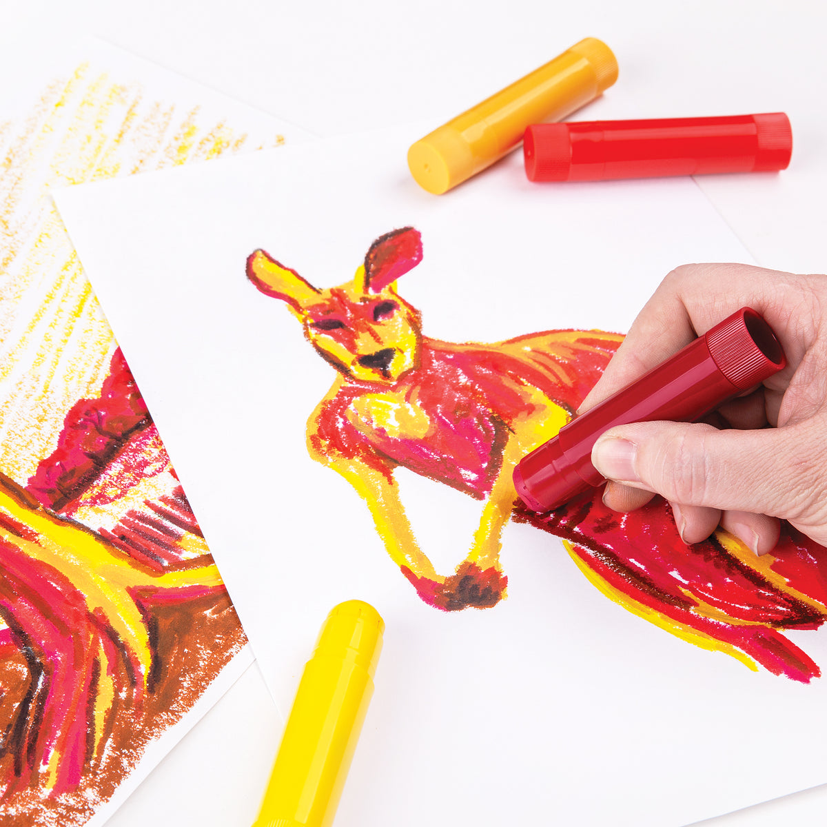 A hand holds a red Ochre Slick, actively colouring the body of an outlined kangaroo drawing in warm red and pink tones. A capped yellow stick lies below, with another yellow and red stick pair resting above. A partly coloured tree and sunburst landscape sits at left.