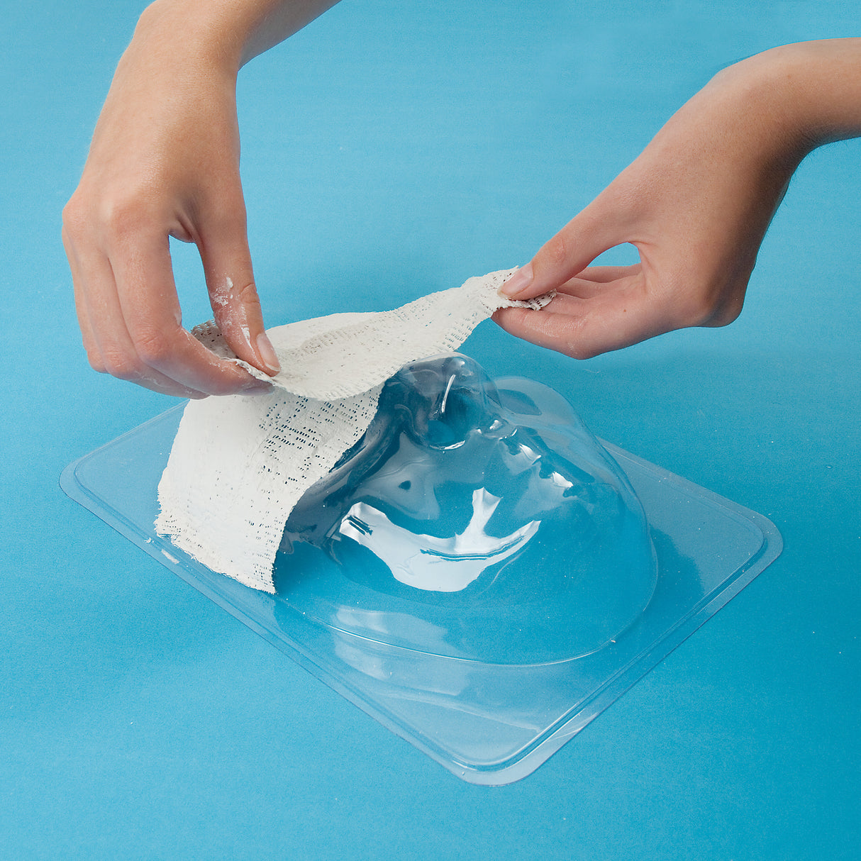 Two hands apply textured plaster bandage strips onto a transparent plastic male face mould positioned on a clear tray against a bright blue background, demonstrating the mask-making process using the Mask Mould Male to create a sculptural face cast with defined facial features.