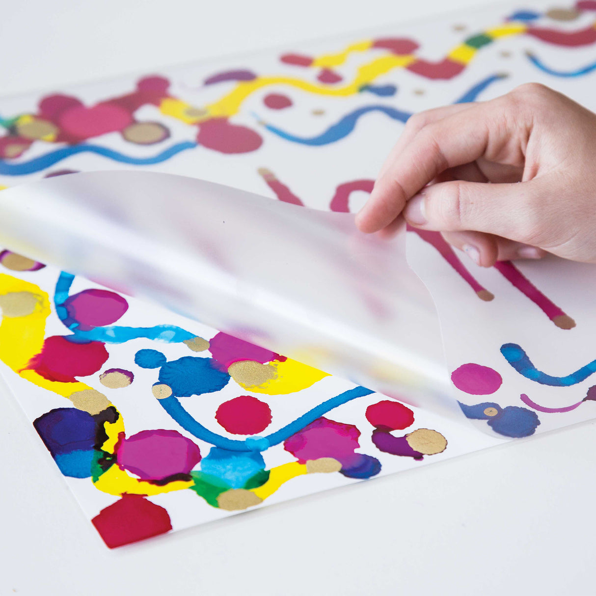 A hand lifts a translucent Zart Laminating Pouch over a colourful abstract artwork featuring bright pink, blue, yellow and gold paint marks. The frosted pouch surface softly blurs the artwork beneath while showing the flexible plastic texture and smooth protective finish.