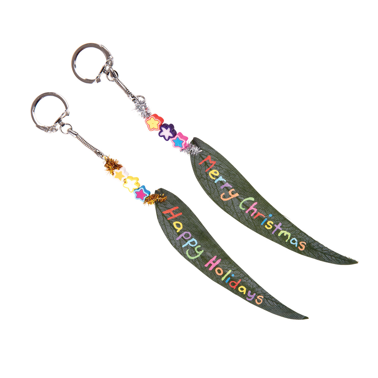 Two keychains crafted from Zart Gum Leaves, each leaf hand-lettered in colourful bubble writing reading "Merry Christmas" and "Happy Holidays". Star and flower-shaped beads plus tinsel sit above each leaf, attached to silver keyring clasps via a small chain link.