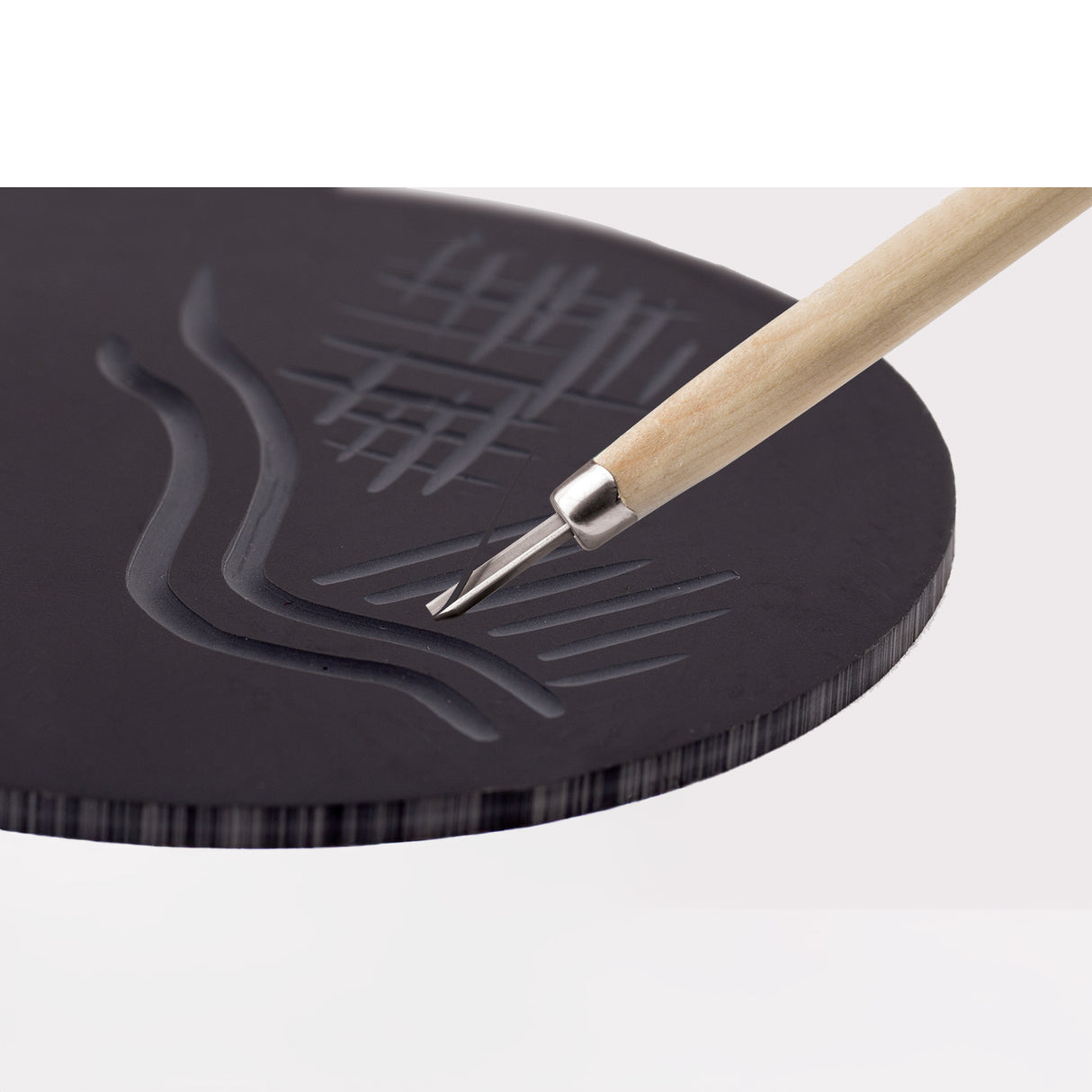 Close-up of a Mastercut Print Lino Round block being carved with a V-shaped lino cutting tool. The dark grey lino surface shows carved grooves, curved lines and textured marks. The tool’s wooden handle and metal blade are positioned mid-cut, demonstrating carving detail.