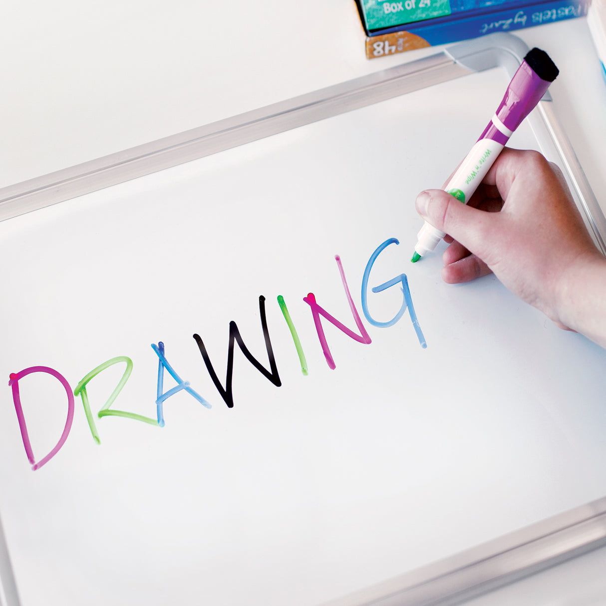 A close-up of a Zart magnetic whiteboard in use, with a hand holding a purple-capped Write 'n' Wipe marker. The word "DRAWING" is written across the surface in rainbow colours. A box of Pastels by Zart, "Box of 24", sits at the top edge.
