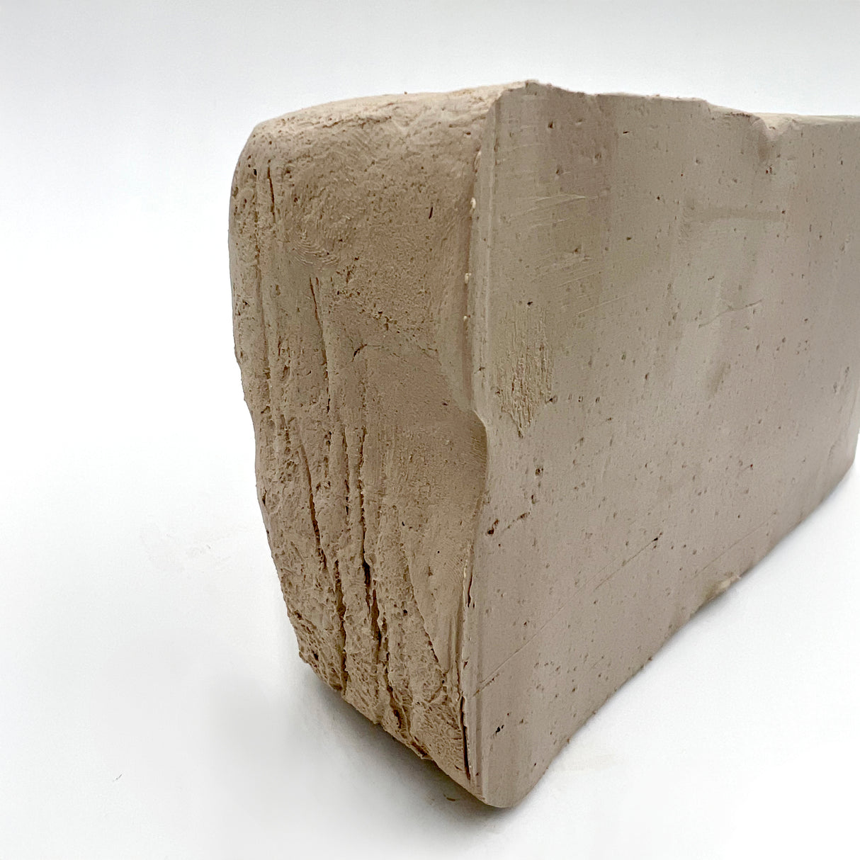 Block of Northcote Stoneware Clay displaying its natural beige-grey colour with darker brown speckles throughout. The rectangular clay block shows textured surfaces with visible tool marks and a smooth cut face, demonstrating the clay's workable consistency and organic appearance on a white background.