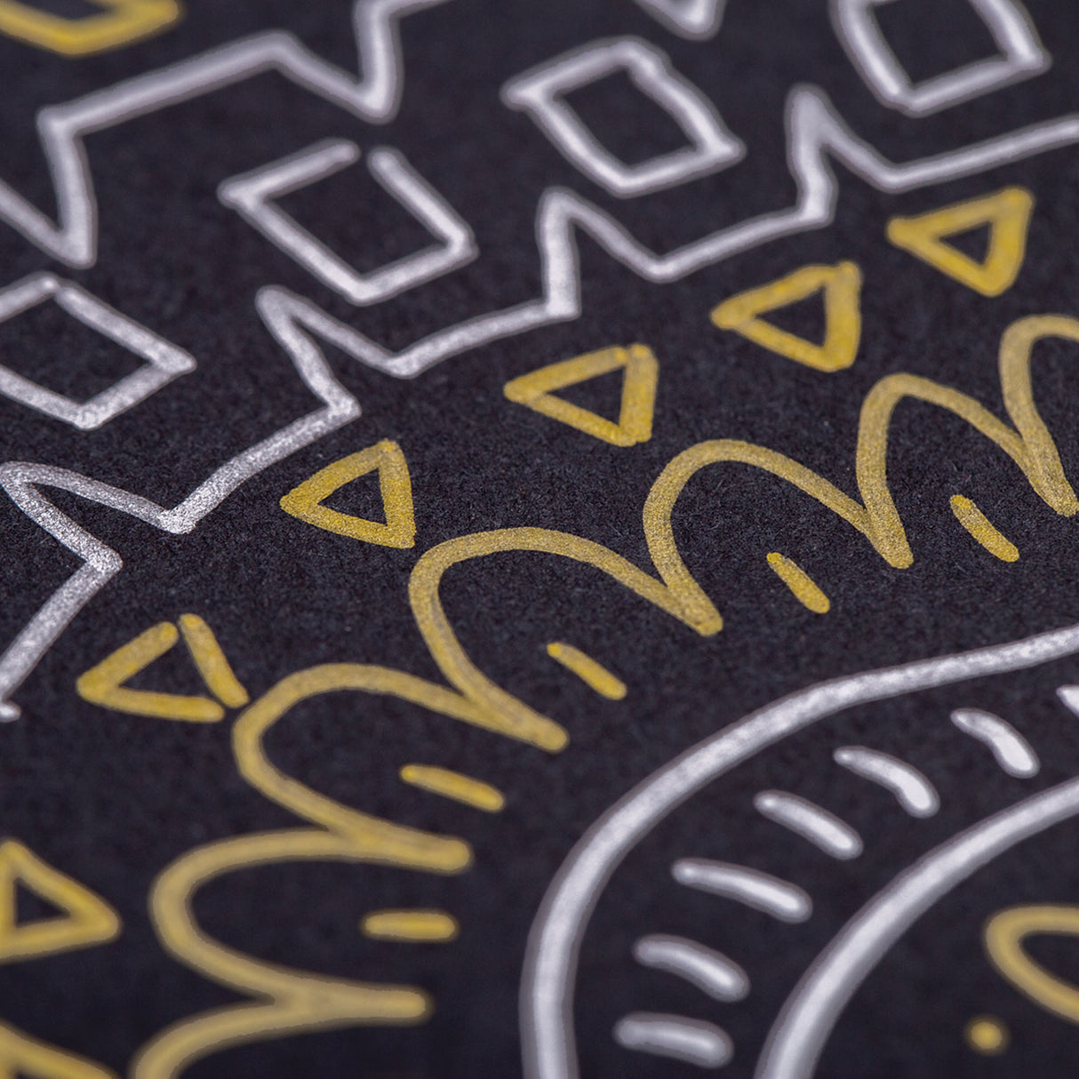 Close-up of metallic artwork on black paper created with Zart Multi Markers showing gold triangular shapes and curved lines alongside silver geometric patterns including squares and zigzag borders. The opaque, reflective ink demonstrates excellent coverage and vibrant metallic finish on dark surfaces.