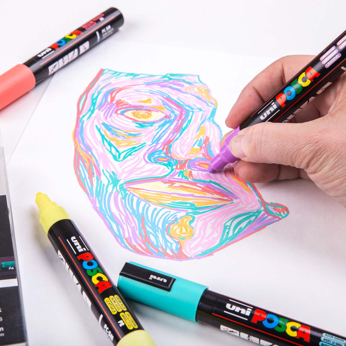 Hand using a purple Uni POSCA pastel marker to draw a stylised multicoloured face made with layered pastel lines. Other POSCA markers lie around the page, showing the vibrant, opaque colours.