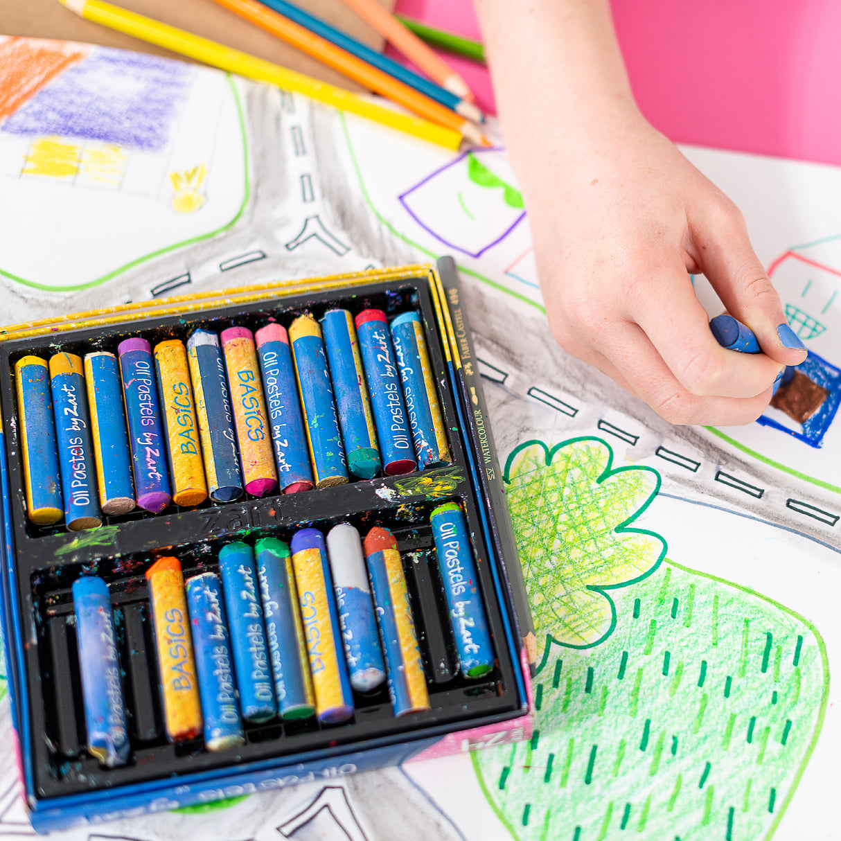 Child’s hand drawing a colourful map scene with oil pastels, beside an open box of Zart oil pastels, showing bright colours, layered marks, and creative use on paper.
