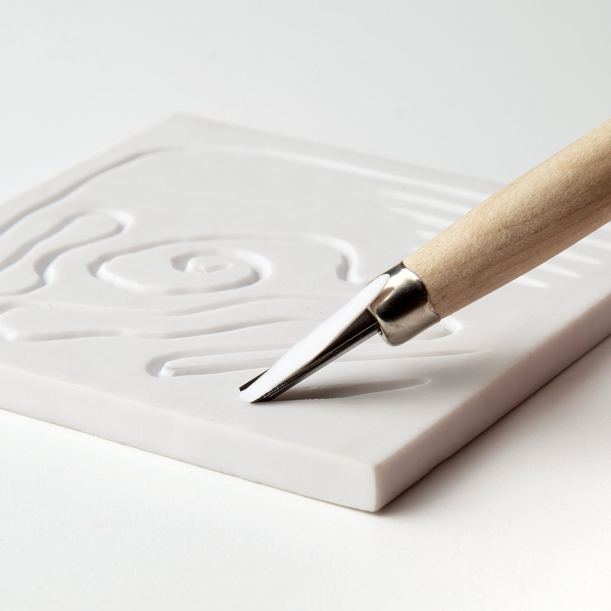 Close-up of V-gouge carving tool cutting into a white soft carving block. Blade creates smooth grooves and curved lines. Wooden handle and metal ferrule visible. Demonstrates precision control and suitability for lino or soft block carving in printmaking and art education settings.