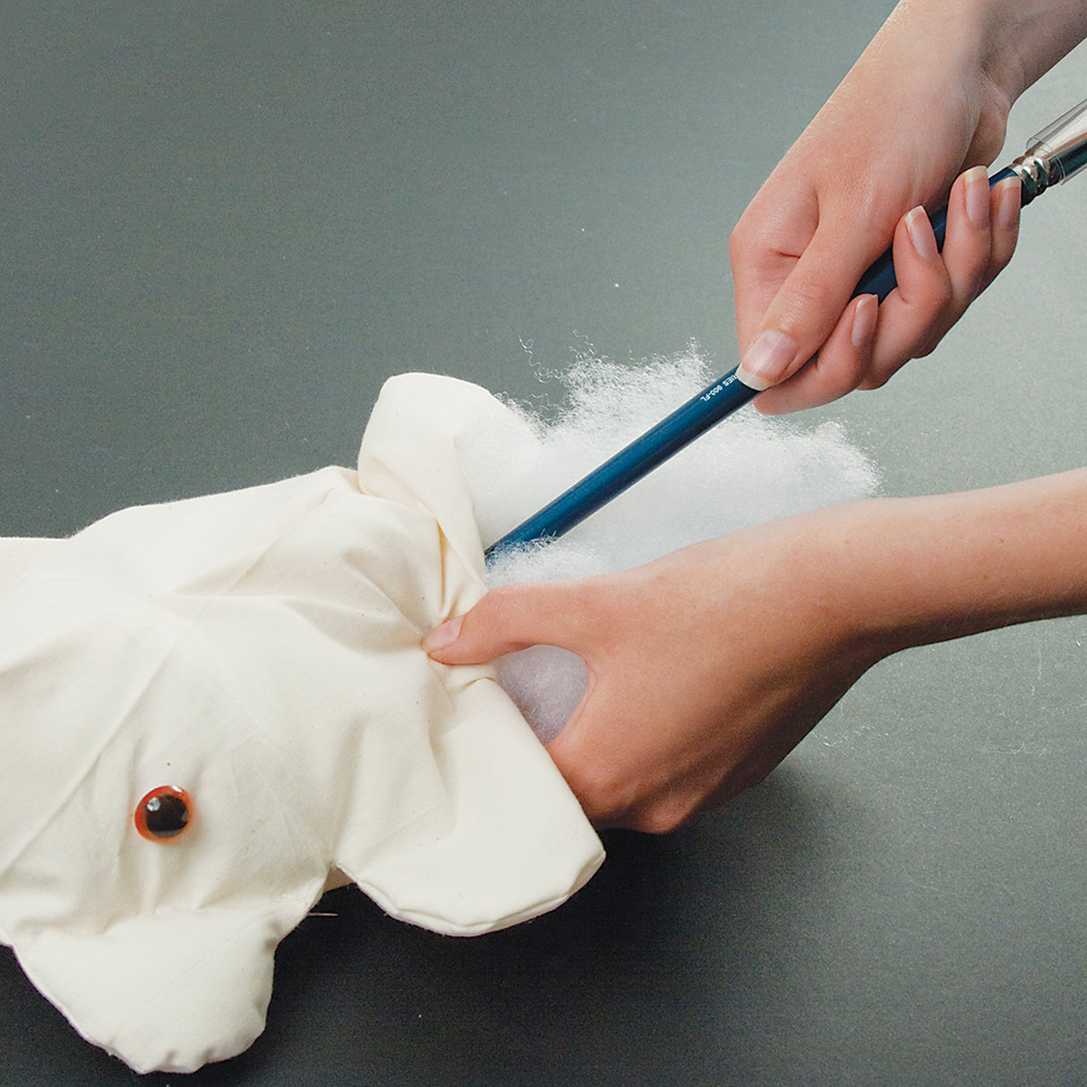 A pair of hands stuffing a cream calico bear with white polyester filling, using a blue-handled tool to push the filling into the body. A safety eye is visible in the foreground. Shown on a dark grey surface, demonstrating the stuffing process.