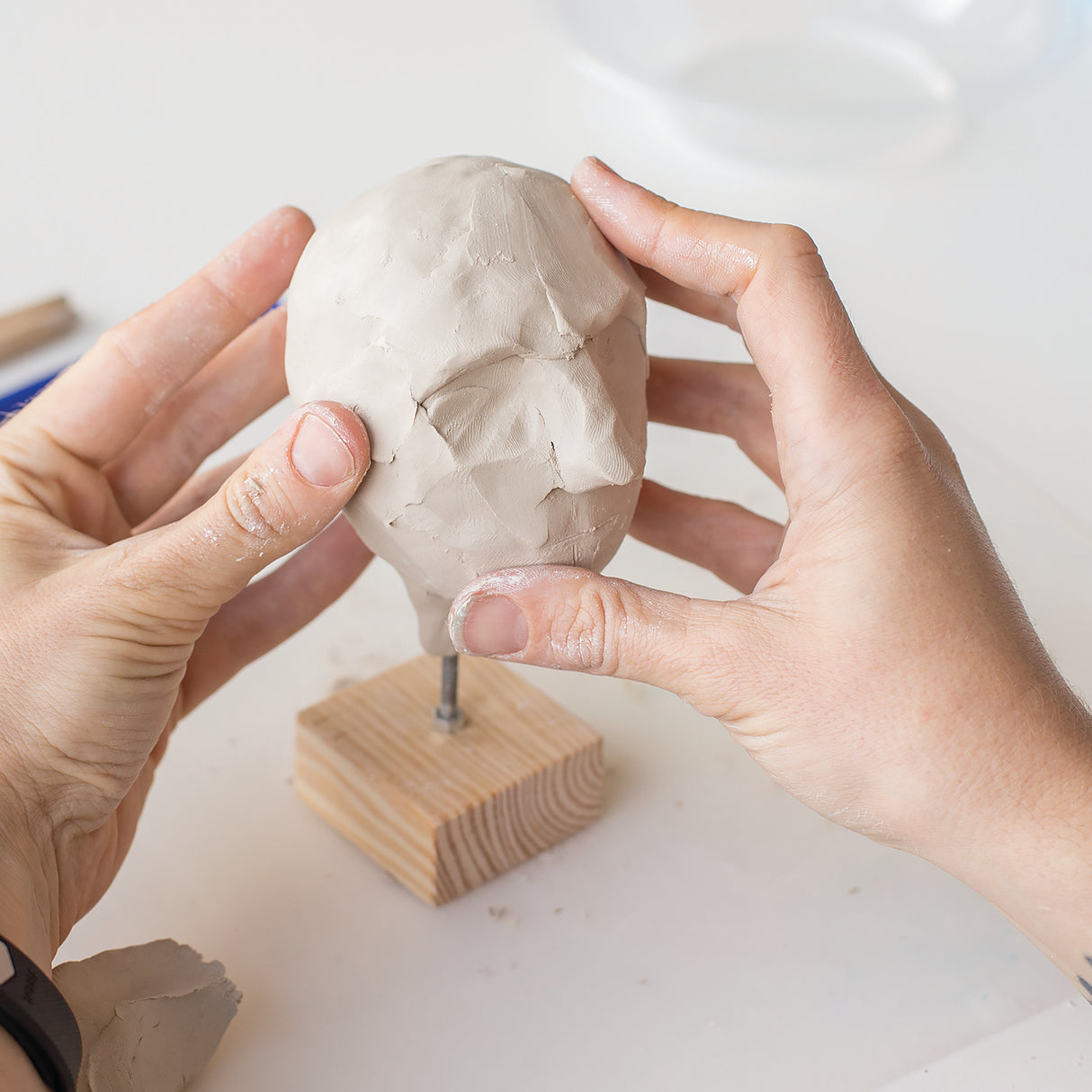 Hands sculpting a grey clay head with visible surface texture and facial features on a wooden armature stand. The malleable Zart Oz Clay shows fingerprint impressions and working marks, demonstrating its smooth, workable consistency ideal for detailed sculptural work and three-dimensional art projects.