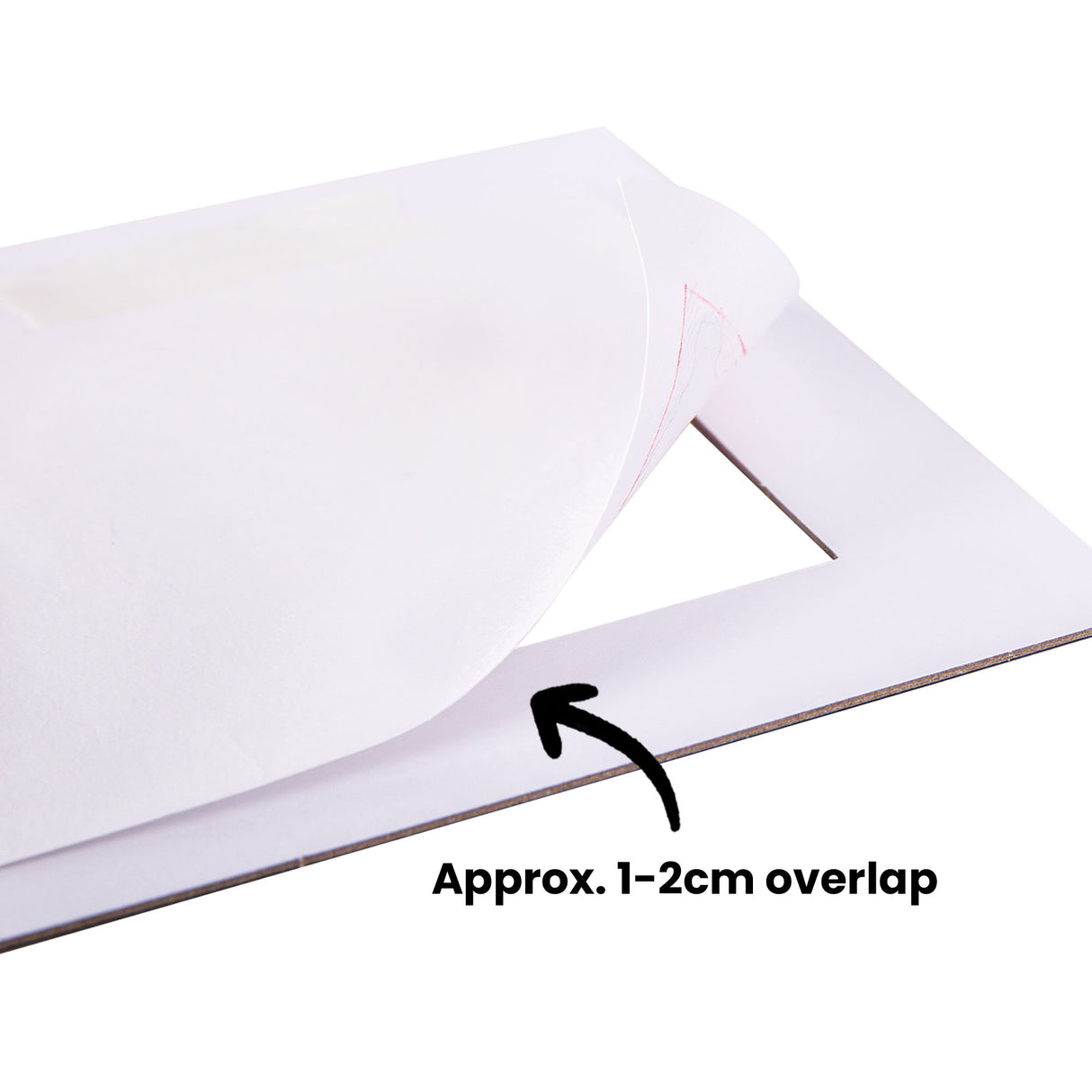 White cardboard mount with rectangular aperture displaying pages curled back to reveal the layered construction. A pink triangle drawing is visible through the opening. Text annotation reads "Approx. 1-2cm overlap" with a black arrow indicating the overlap between mount layers.