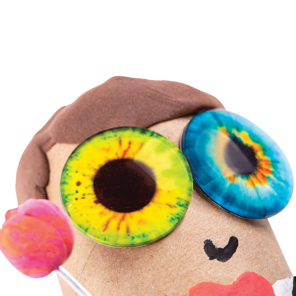 Cardboard character head with textured brown hair featuring two large dome-shaped Creature Eyes in vibrant colours: one with yellow and green iris patterns, another with blue and orange detailing. Pink pom-pom ear, black drawn smile, and red mouth accent complete the playful sculptural creation.