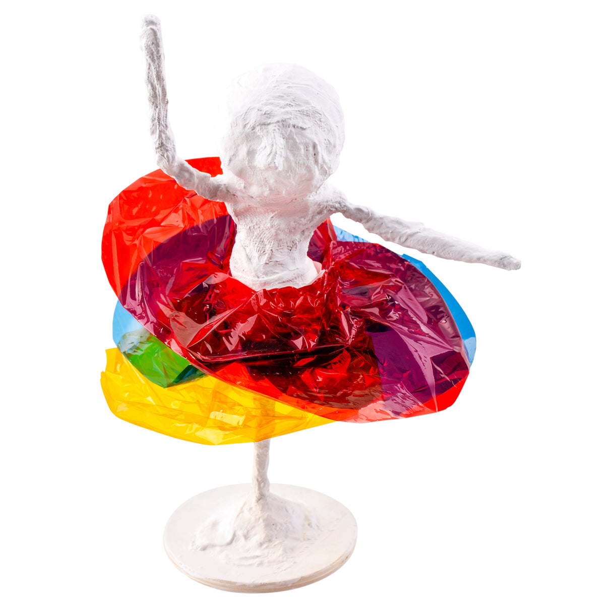 Small sculptural figure with outstretched arms mounted on a round base, wearing a layered skirt made from red, yellow, green and blue cellophane. The translucent material creates a bright, dimensional effect resembling a colourful tutu.