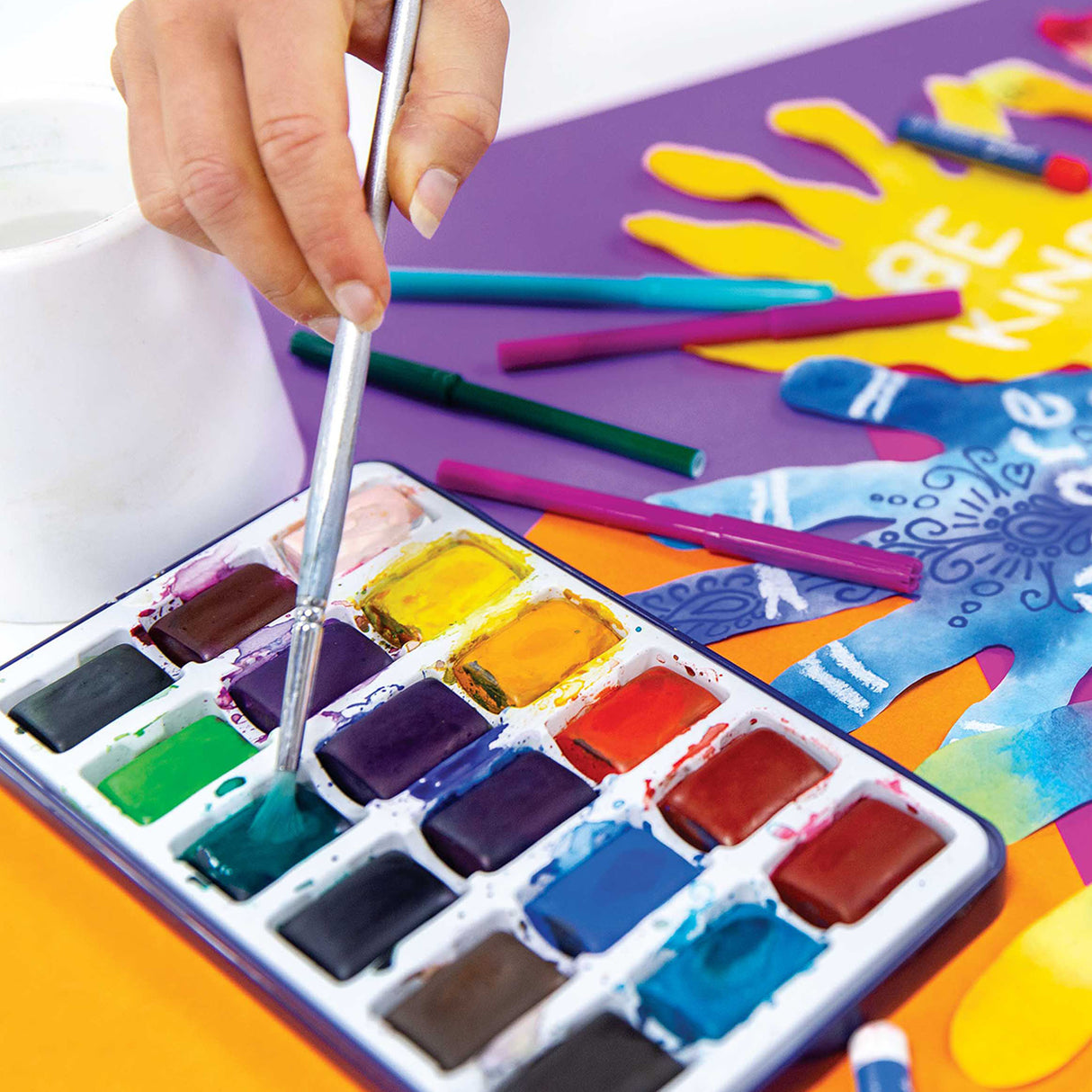 A hand holds a paintbrush dipping into a well-used watercolour palette containing vibrant colours including greens, purples, yellows, oranges, reds and blues. Colourful art supplies scatter the workspace including gel crayons in teal, magenta and purple, alongside decorative paper cutouts featuring a yellow sunshine design and blue patterned elements on a purple background.