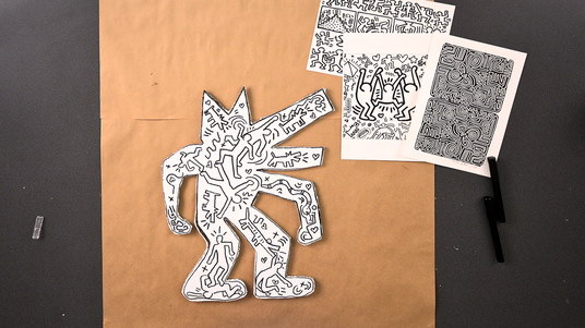 A cut-out cardboard figure covered in bold black doodles — dancing figures, hearts, stars and graphic symbols — displayed against brown kraft paper. Printed Keith Haring reference sheets are visible alongside a black marker. Zart Education On Demand lesson activity: Cardboard Doodle Character Inspired by Keith Haring.