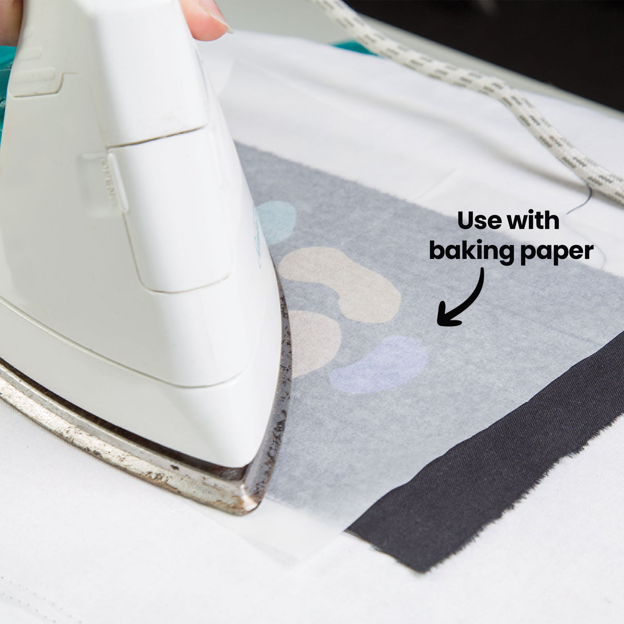 A close-up of a white iron pressing over baking paper laid on black fabric, with pastel-toned glitter shapes from Zart Glitter Iron-On Sheets visible beneath. The annotation 'Use with baking paper' points to the paper, showing the correct method for applying the sheets.