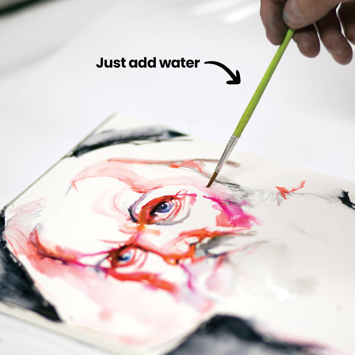 A hand using a wet brush to activate and blend Artline Liquid Crayons on a portrait drawing, creating soft watercolour washes in red and pink tones. An annotation reads "Just add water," demonstrating the product's water-reactive properties.