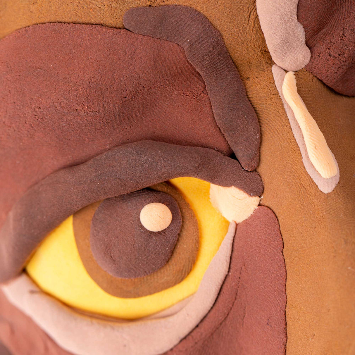 Close-up of a sculpted face detail created with Paper Magiclay in earth colours, showing layered terracotta, brown, ochre, pink and cream tones. The textured eye features concentric circles with a yellow iris and brown pupil, demonstrating the clay's blendability and matte, paper-like finish with visible surface texture.
