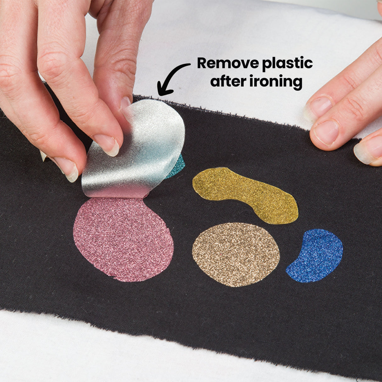 Two hands peel a silvery plastic backing away from a pink glittered shape on black fabric, revealing gold, tan and blue Zart Glitter Iron-On Sheets shapes alongside. The annotation 'Remove plastic after ironing' points to the backing, showing the final step in application.