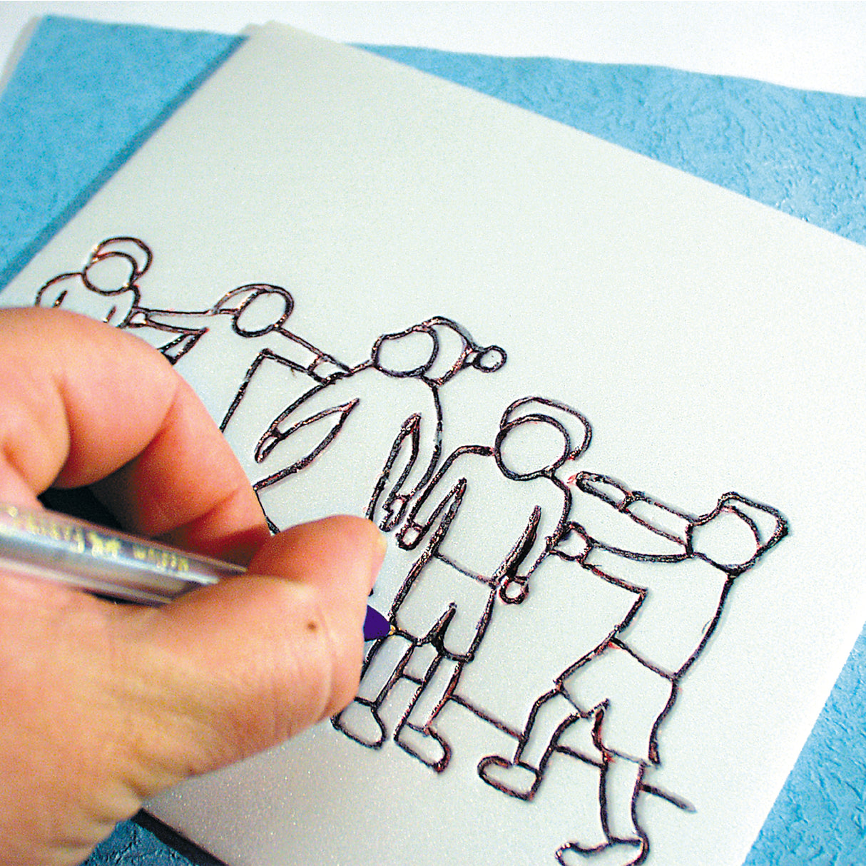 Close-up of a hand using a pen to draw simplified figures onto a print foam sheet. The figures show people standing and moving in different poses. The foam surface is pale and smooth, with bold black outlines forming the design before etching and printing.