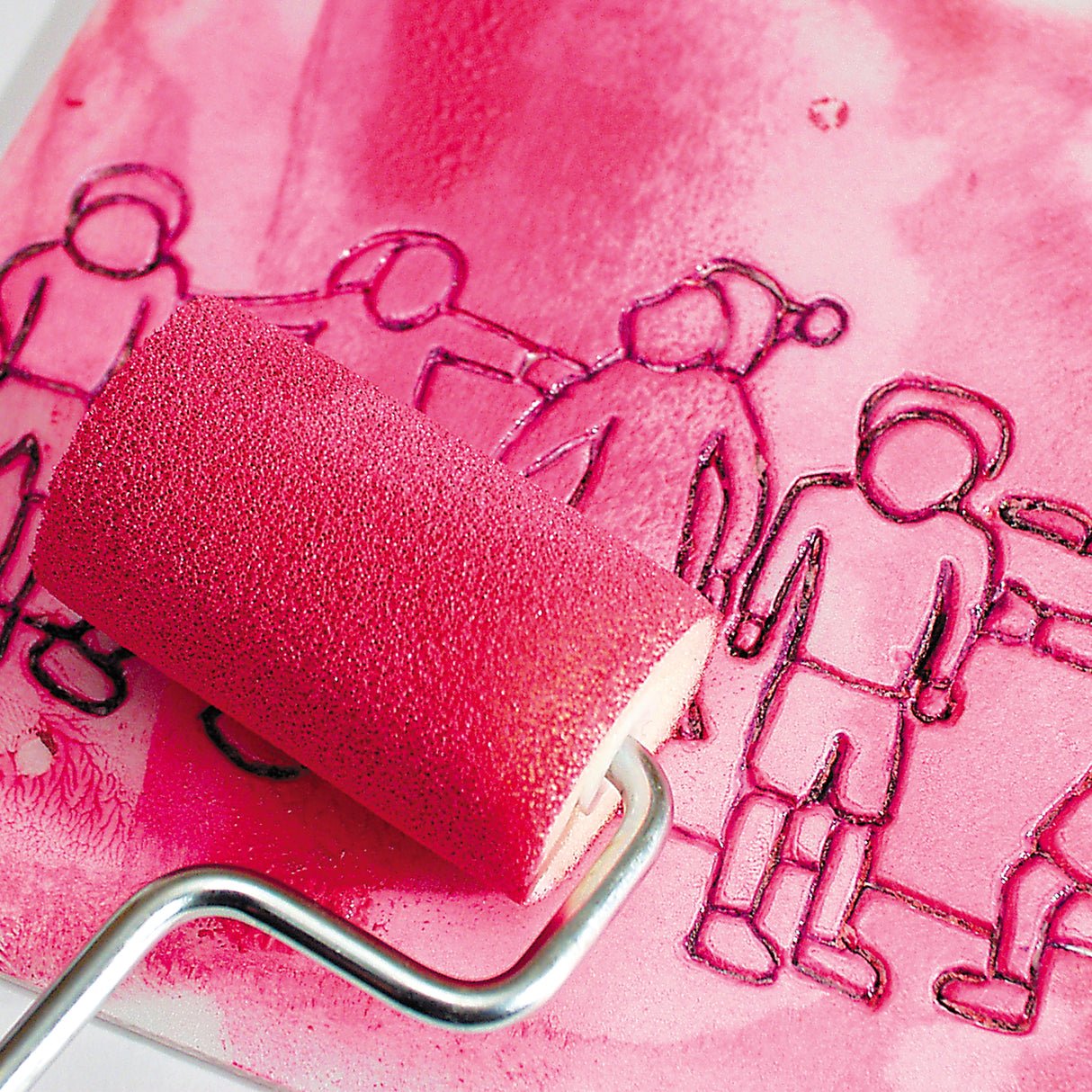 A pink ink roller is pressed onto a print foam plate covered in pink ink. The foam plate shows outlined figures of people, with raised and indented areas visible through the ink layer. The image demonstrates inking the print foam surface before transferring a print.
