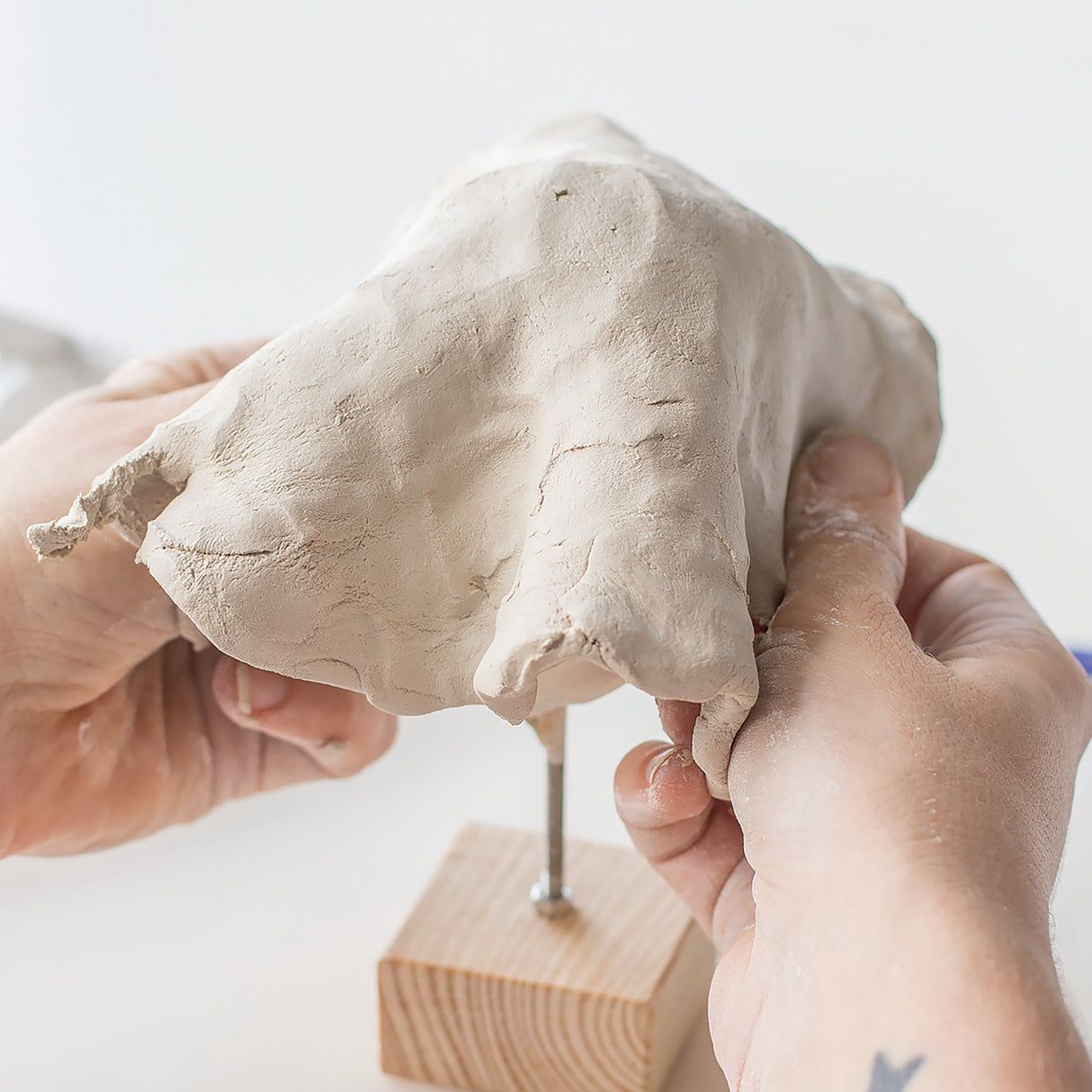 Two hands hold a piece of soft, pliable Zart Oz Clay above a wooden armature base with wire support. The cream-coloured clay shows a smooth, workable texture with natural surface variations and fingerprints, demonstrating its sculptural properties ideal for modelling and hand-building projects in educational and studio settings.