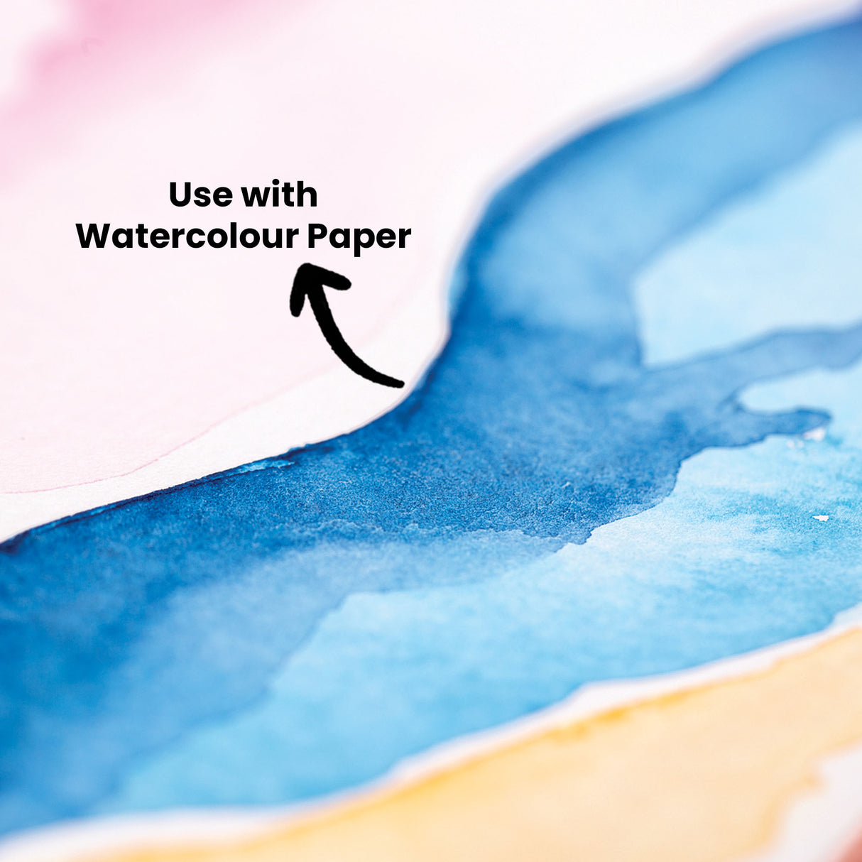 Close-up of watercolour painting showing vibrant blue washes blending into soft pink and warm yellow tones on textured paper. Text annotation reads "Use with Watercolour Paper" with an arrow pointing to the blue painted area, demonstrating the Primecolours Watercolour Palette's colour blending capabilities.