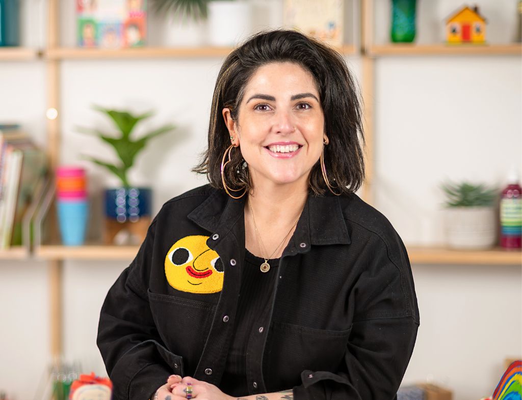 Woman wearing black denim jacket with cheerful yellow emoji patch featuring sunglasses and red smile, standing in bright art classroom with colourful student artwork and craft supplies displayed on wooden shelves behind her, embodying creative learning environment for Art & Literacy education.