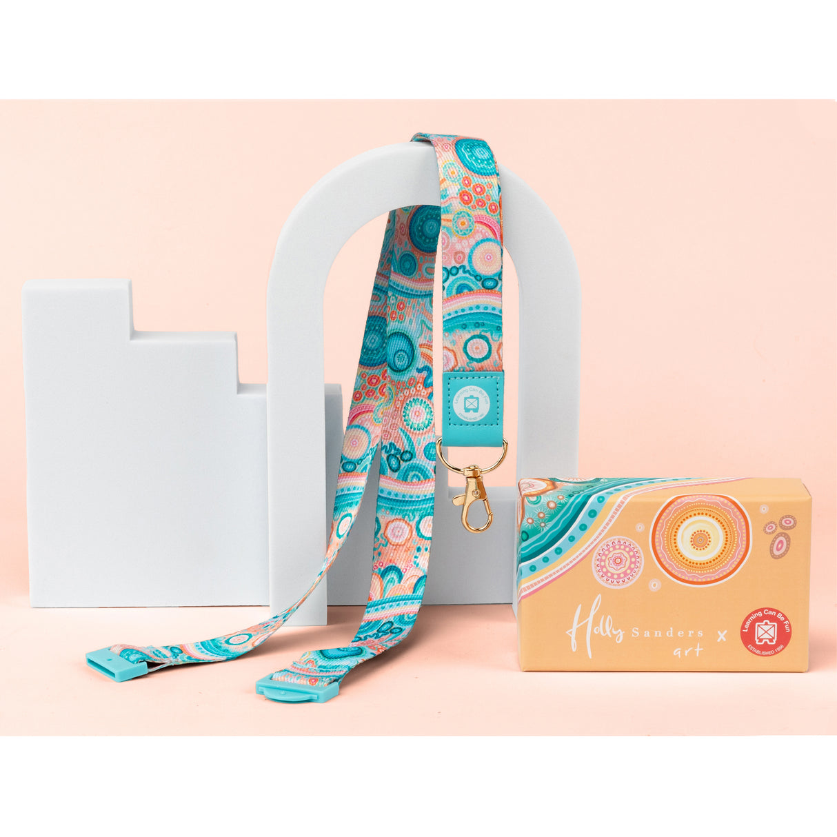 Holly Sanders x art lanyard with gold swivel clip, featuring vibrant Indigenous Australian designs in turquoise, coral, and peach tones with circular dot patterns and curved motifs. The lanyard is displayed draped over a white sculptural display alongside its matching gift box showing concentric circle artwork.