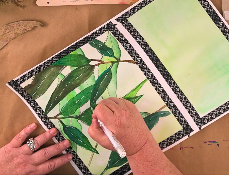 Zart Education PL Online Exploring Watercolour and Ink — A person adds fine white pen details to a watercolour painting of eucalyptus leaves. The artwork is bordered with patterned tape, and a second sheet with a light green wash sits beside it. Hands, brushes, and natural leaf references rest on the brown paper workspace, highlighting a focused botanical illustration process.