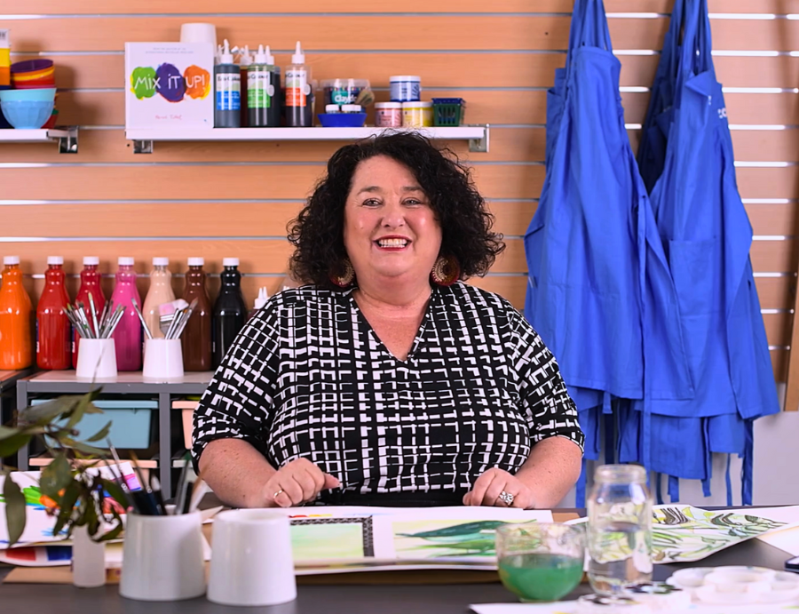 Smiling educator seated at a workspace with watercolour artwork samples displayed on the table. Behind her, shelves display colourful paint bottles, brushes in containers, and a MIX IT UP! sign. Blue aprons hang on the right side of the peach-coloured slatwall backdrop.