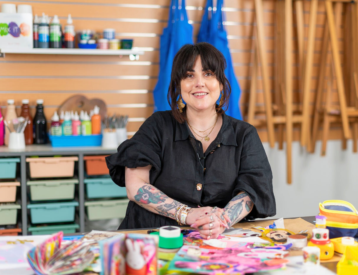 Woman with dark hair and tattooed arms wearing black shirt sits at table covered with colourful collage supplies including paper, adhesives, and craft materials. Behind her are shelves displaying art supplies, paint bottles, brushes in containers, and blue aprons hanging on a wooden slatwall in a bright creative studio space.
