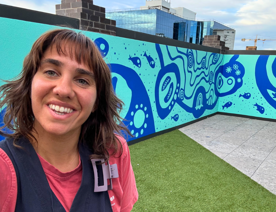 Kirli Saunders smiling in front of a vibrant blue mural featuring flowing, symbolic shapes on an outdoor wall, with urban buildings in the background. From Zart Education Live Online with Kirli Saunders – images provided by Lilli Paton on behalf of the artist.