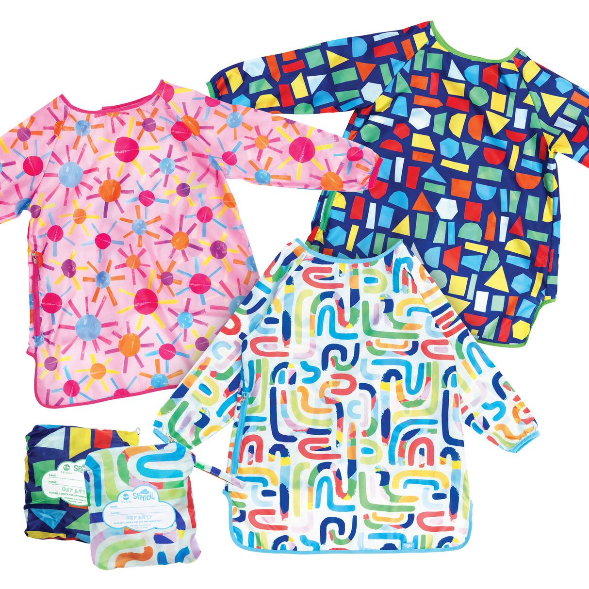 Three colourful Spencil Art Smocks displayed flat, featuring vibrant patterns: pink with multicoloured starburst design, navy with geometric shapes in yellow, red, green and blue, and white with abstract curved lines in rainbow colours. Small folded smock with packaging visible in corner.