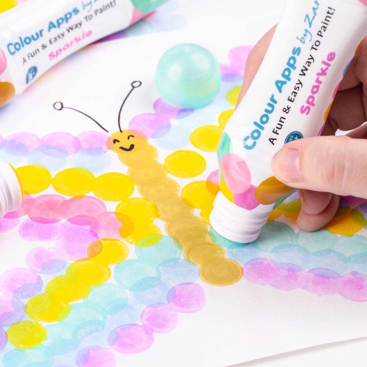 Hand using a Zart Colour Apps Sparkle bottle to stamp overlapping pastel dots onto paper. Yellow, pink, blue and green circles form a butterfly shape, with a loose cap nearby.