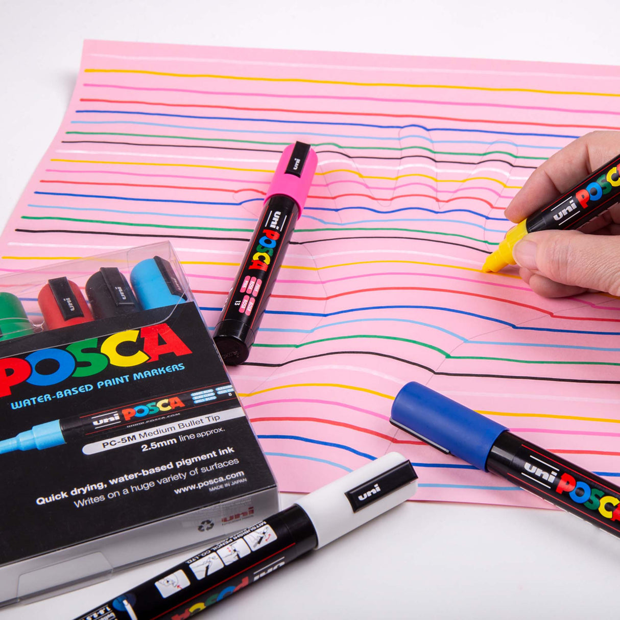 Hand using a yellow POSCA marker to draw curved coloured lines on pink paper for an optical-illusion style hand artwork. POSCA markers and packaging sit around the page.