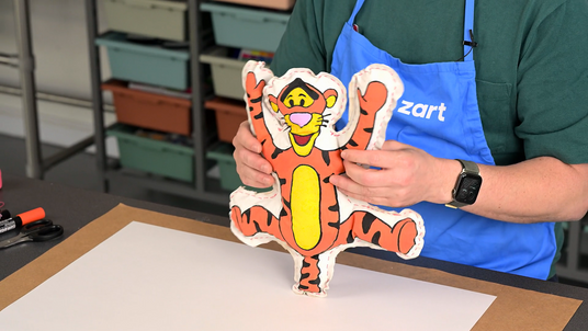 Zart educator holds up a completed soft sculpture of Tigger from Winnie-the-Pooh, created as part of a Zart Education On Demand lesson activity. The fabric figure is painted in bold orange, black, and yellow with a cheerful expression, and features visible red running-stitch seams around the cut-out calico form.