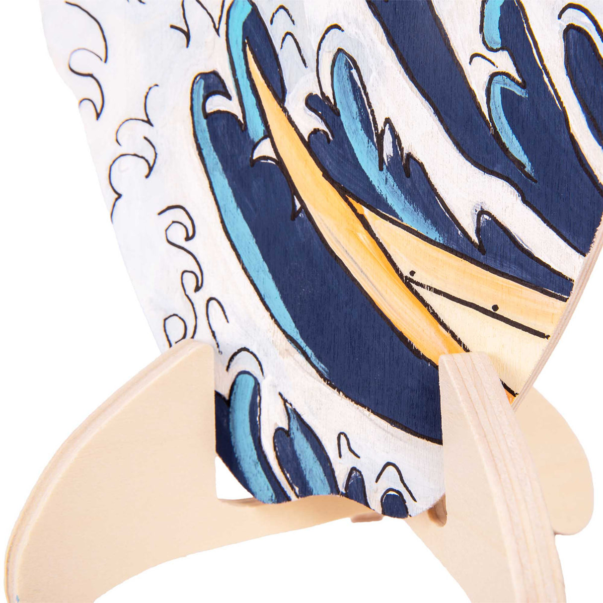 Close-up of a student-painted surfboard-shaped plywood artwork seated in a wooden stand. The artwork features layered blue waves with white highlights, echoing The Great Wave style, with visible brushstrokes and curved foam details near the base.