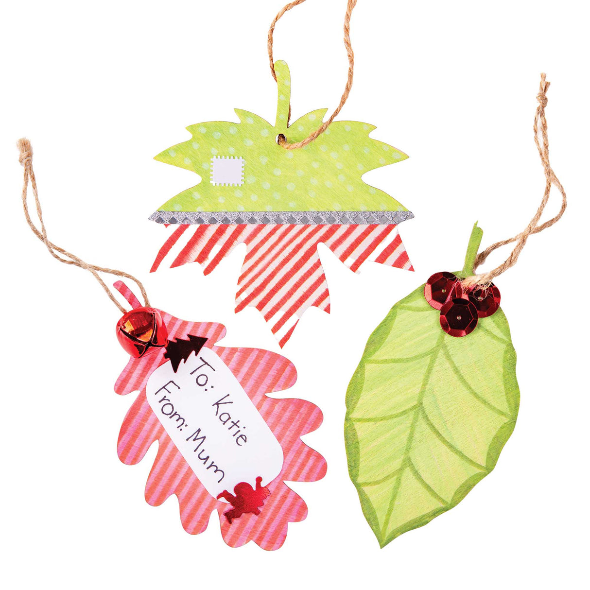 Three Zart Wooden Leaf Adornments hung on jute string, decorated as gift tag samples. One maple leaf shows green polka-dot and red striped washi tape with a paper stamp motif; another has pink stripes, a red bell and a tag reading "To: Katie From: Mum"; the third is a plain green leaf with red sequin berries.