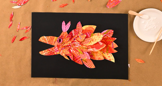 Bright creature collage made from layered gel plate printed paper shapes in shades of red, orange and pink, arranged on black card to form a whimsical animal. Glue and collage materials surround the artwork (Zart Education On Demand Gel Plate Creature Collage activity).