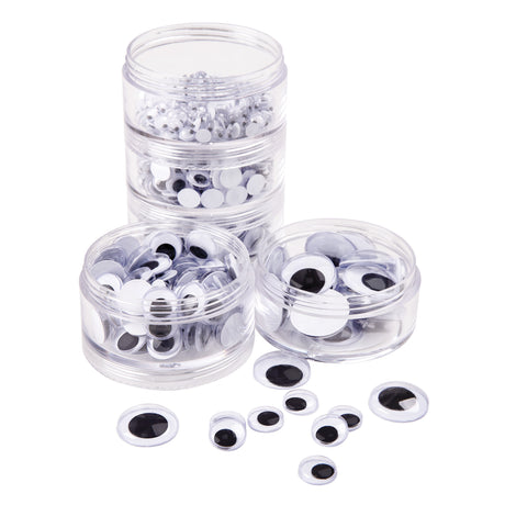 Zart Joggle Eyes shown in three clear round tubs of mixed sizes, with one tub stacked three-high behind two single tubs at the front. Loose Joggle Eyes are scattered beside the tubs, each a round black pupil disc that slides inside a clear plastic dome on a white base.