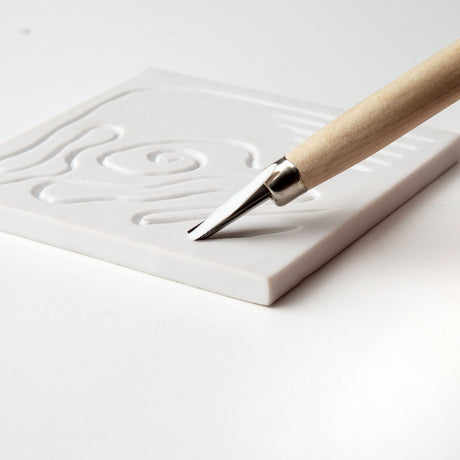 Close-up of a wooden-handled carving tool cutting into a white soft cut block. The metal blade creates smooth recessed lines forming curved shapes. The block has a matte surface and straight, clean edges, resting on a white background.