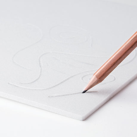 Close-up photo of a sharpened wooden pencil drawing into a white print foam sheet. A faint indented line is visible where the pencil tip presses into the soft surface, with a lightly sketched outline design in the background. The image demonstrates marking and preparing print foam for printing.