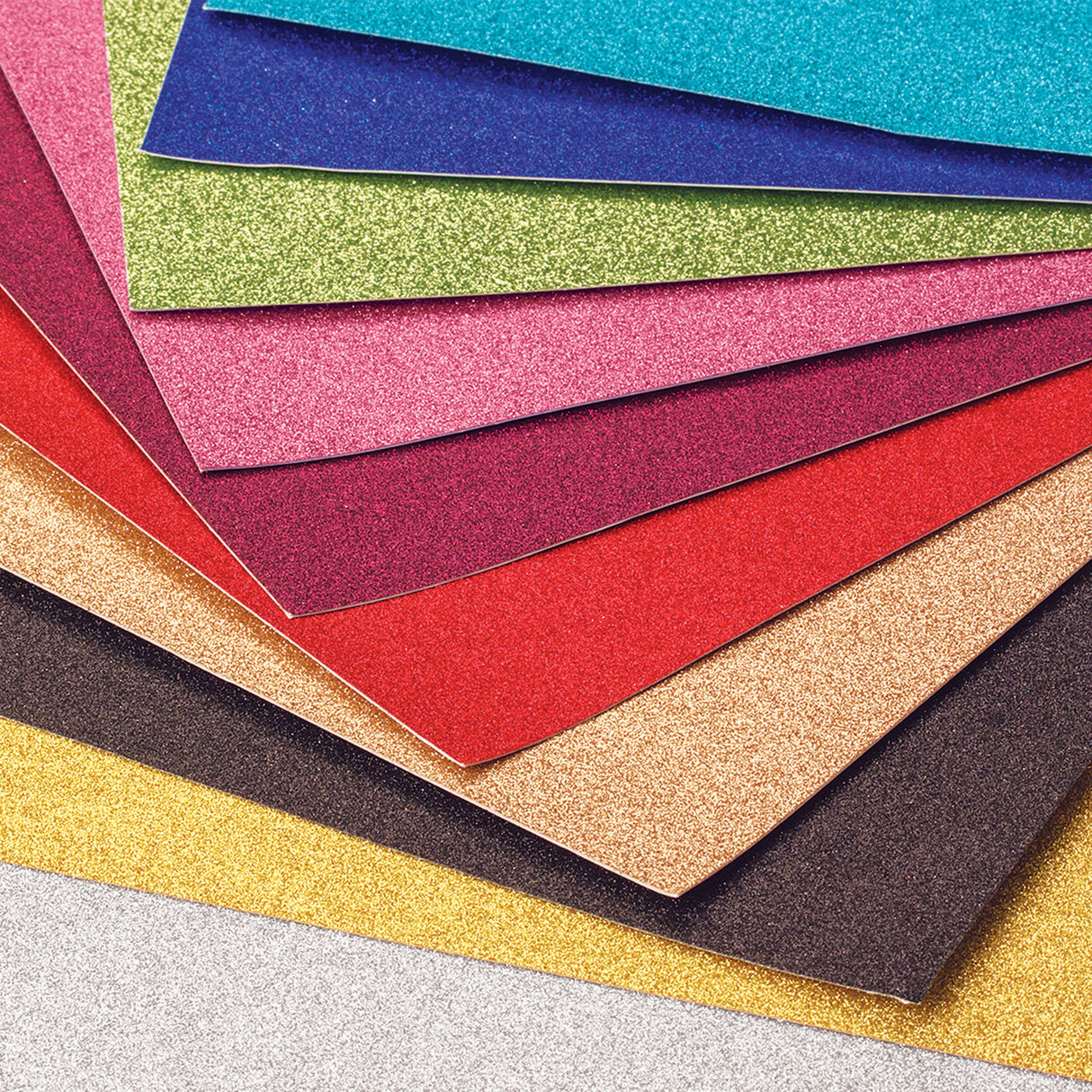 Zart Glitter Iron-On Sheets fanned out to reveal a vivid range of colours, including pink, teal, blue, green, magenta, red, gold, bronze, black, yellow and silver. Each sheet has a fine, densely packed glitter finish with a soft sheen, showcasing the full assorted colour palette.