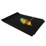 A stack of black-coated Scratch Rainbow Scraper Card sheets, the top sheet partly scratched to reveal a rainbow scribble in blue, green, yellow, orange and red. A pointed wooden scraper tool rests on the surface where the black coating has been scratched away.