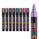 POSCA Metallic Marker set of eight shown with lids on in a row, each cap coloured to match its ink: gold, silver, bronze, blue, green, red, pink and violet. A larger uncapped violet marker sits beside them, showing its rounded metallic nib and black barrel with coloured text.